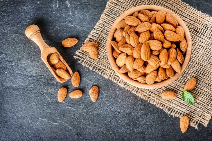 Top view of almonds on dark stone table with wood spoon or scoop. Almond in wooden bowl. Nuts freely laid on dark board.